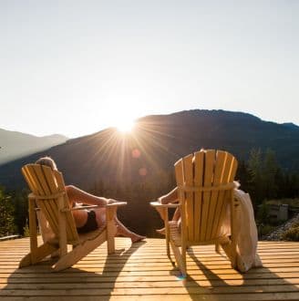 two people sit on adirondack chairs on a deck in hammock haven at sunset looking at the sun setting behind the mountains