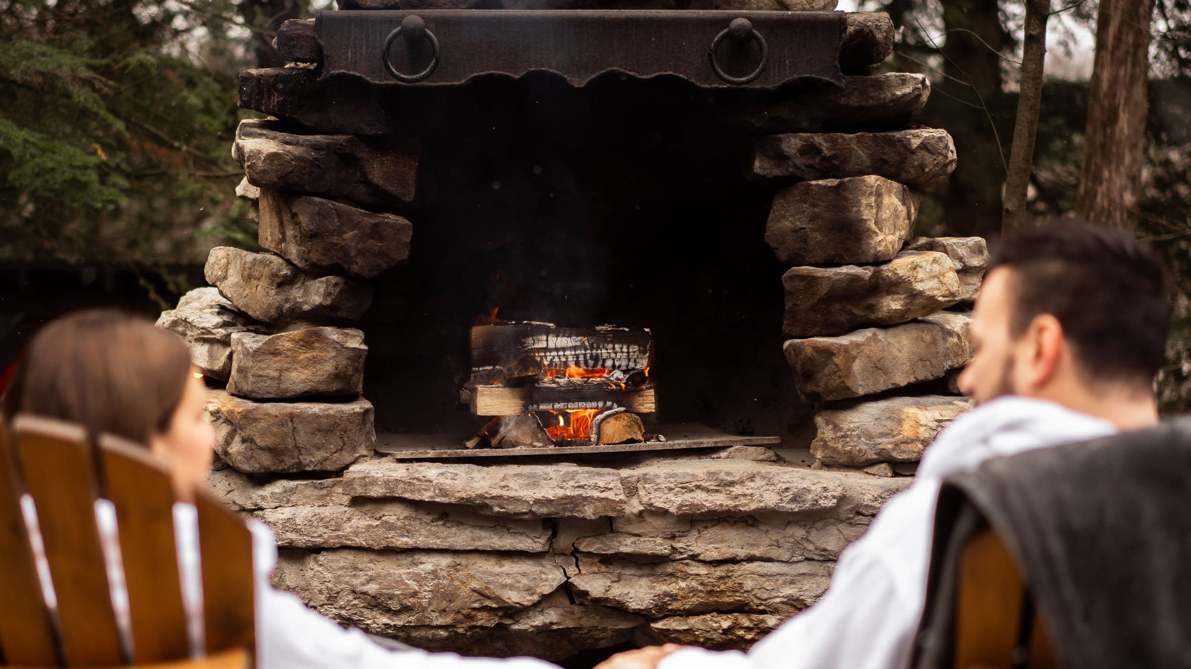 A couple relax in front of an outdoor stone fireplace