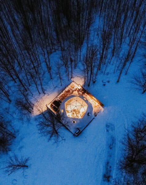 Geodesic dome in the wilderness at dusk, with an aerial view of a forested landscape.