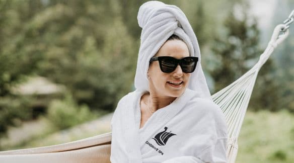 a women in a white spa robe and towel on her head as a turban, smiles with sunglasses on while lounging in a hammock at Scandinave Spa Whistler, mountains in the background