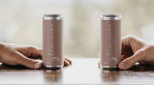 Two hands each hold a can of glimmer dealcoholized sparking wine in the spa bistro at Scandinave spa whistler