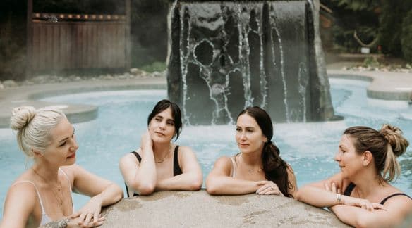 The women of Glimmer relax together in the hot pool at Scandinave Spa Whistler, thermal waterfall in the background