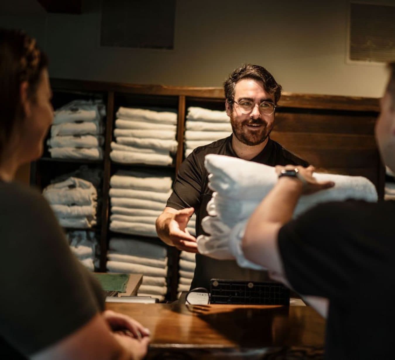 Un agent du service clientèle à l'air sympathique tend des serviettes pliées aux clients à un comptoir d'accueil, avec des étagères remplies de serviettes en arrière-plan.