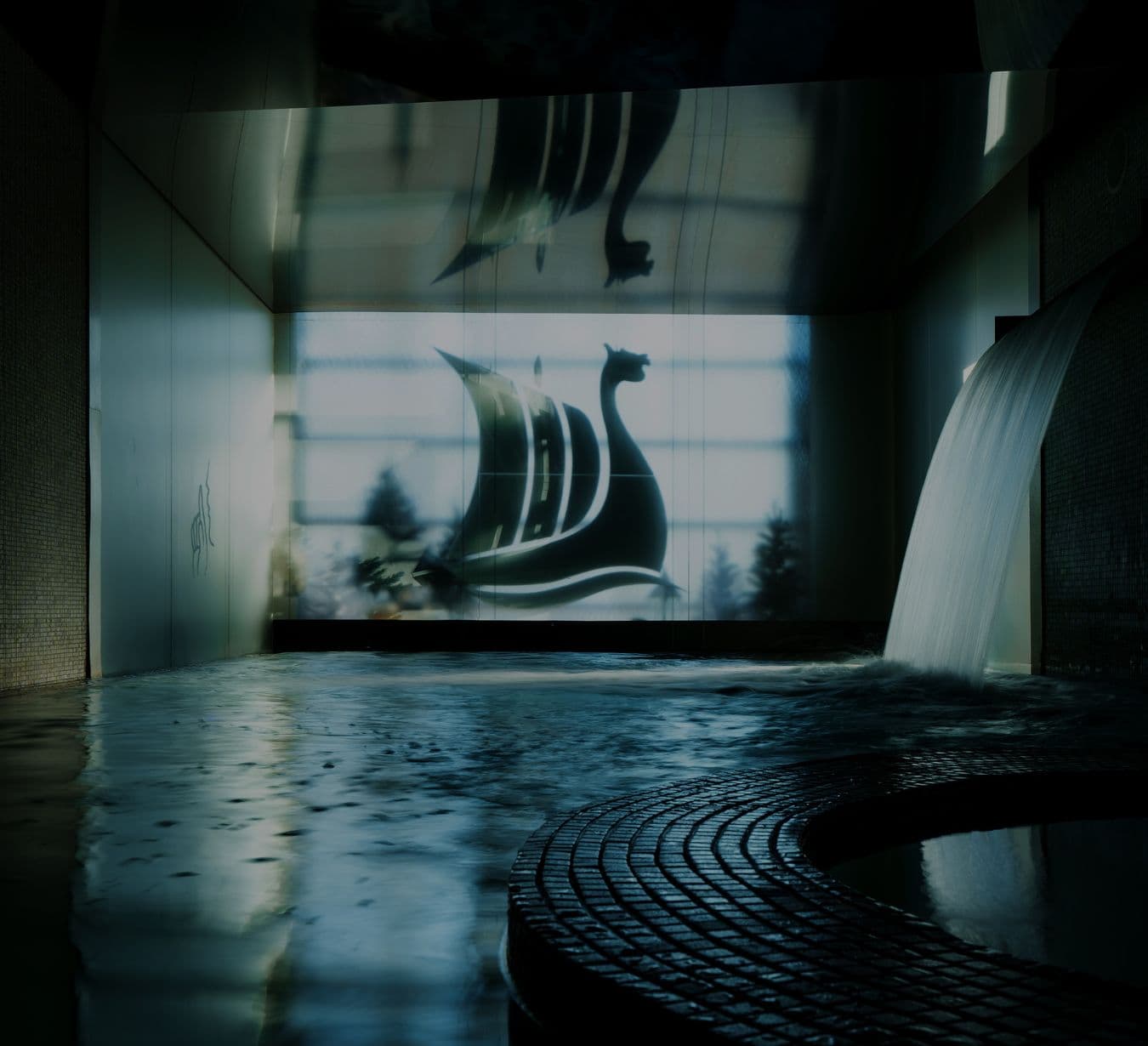 Indoor pool with a thermal waterfall in the foreground. In the background, an illuminated wall displays the Drakkar, the Scandinave Spa logo, designed as a stylized Viking ship, creating a soothing ambiance.