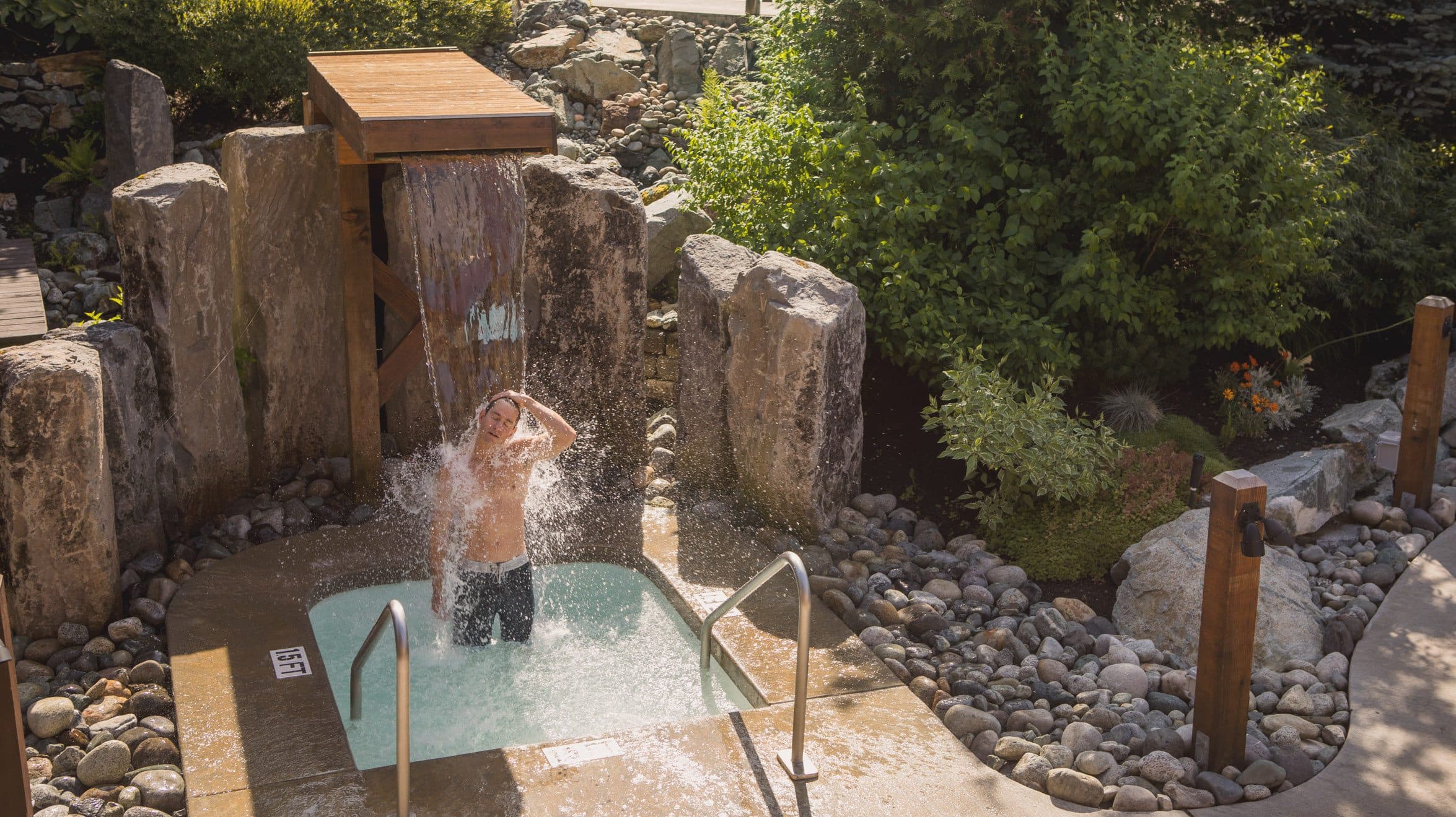 Un homme s'étirant le cou sous la cascade nordique du Scandinave Spa Whistler tout ent profitant des rayons du soleil d'été.