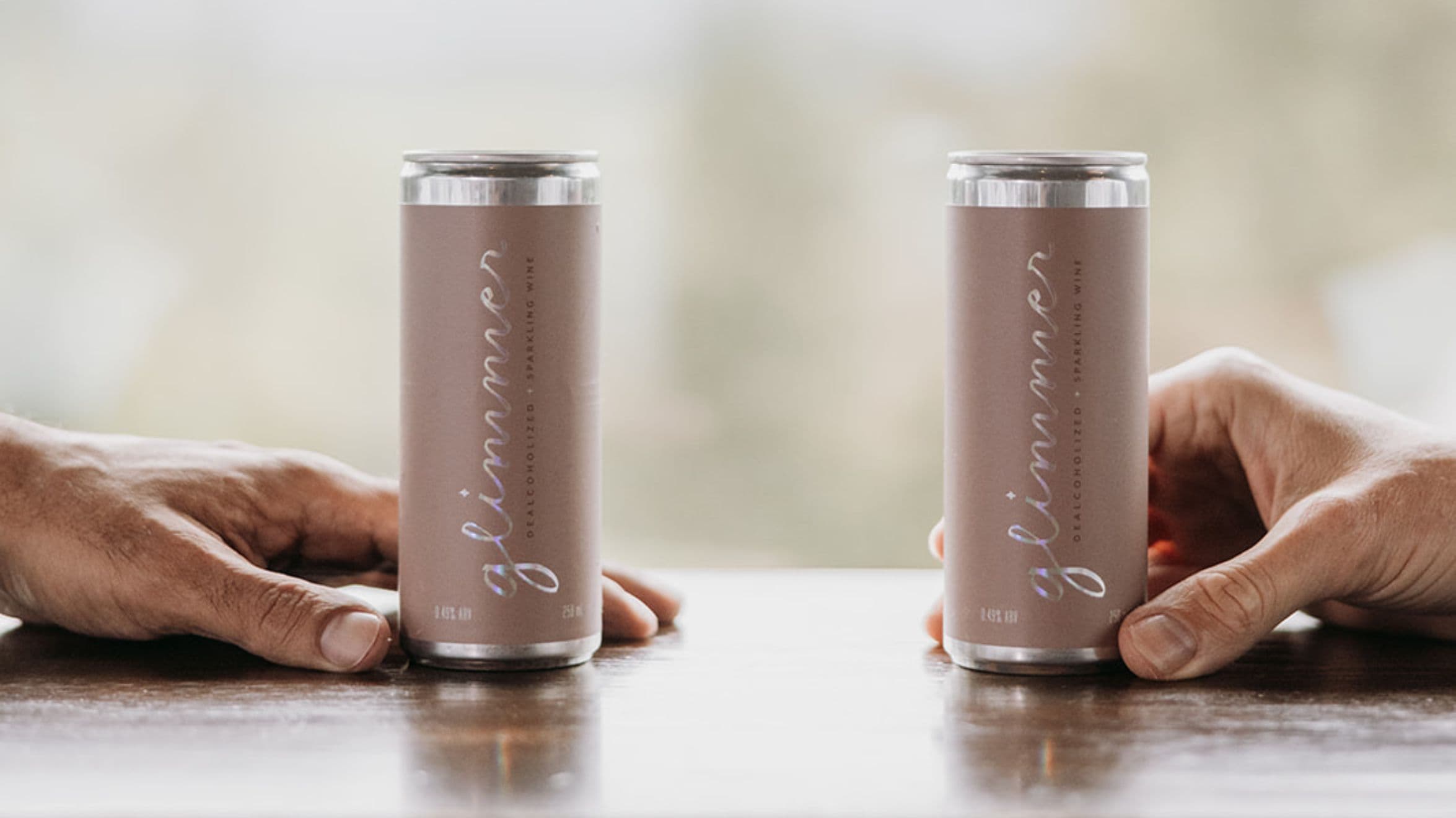 Two hands each hold a can of glimmer dealcoholized sparking wine in the spa bistro at Scandinave Spa Whistler.