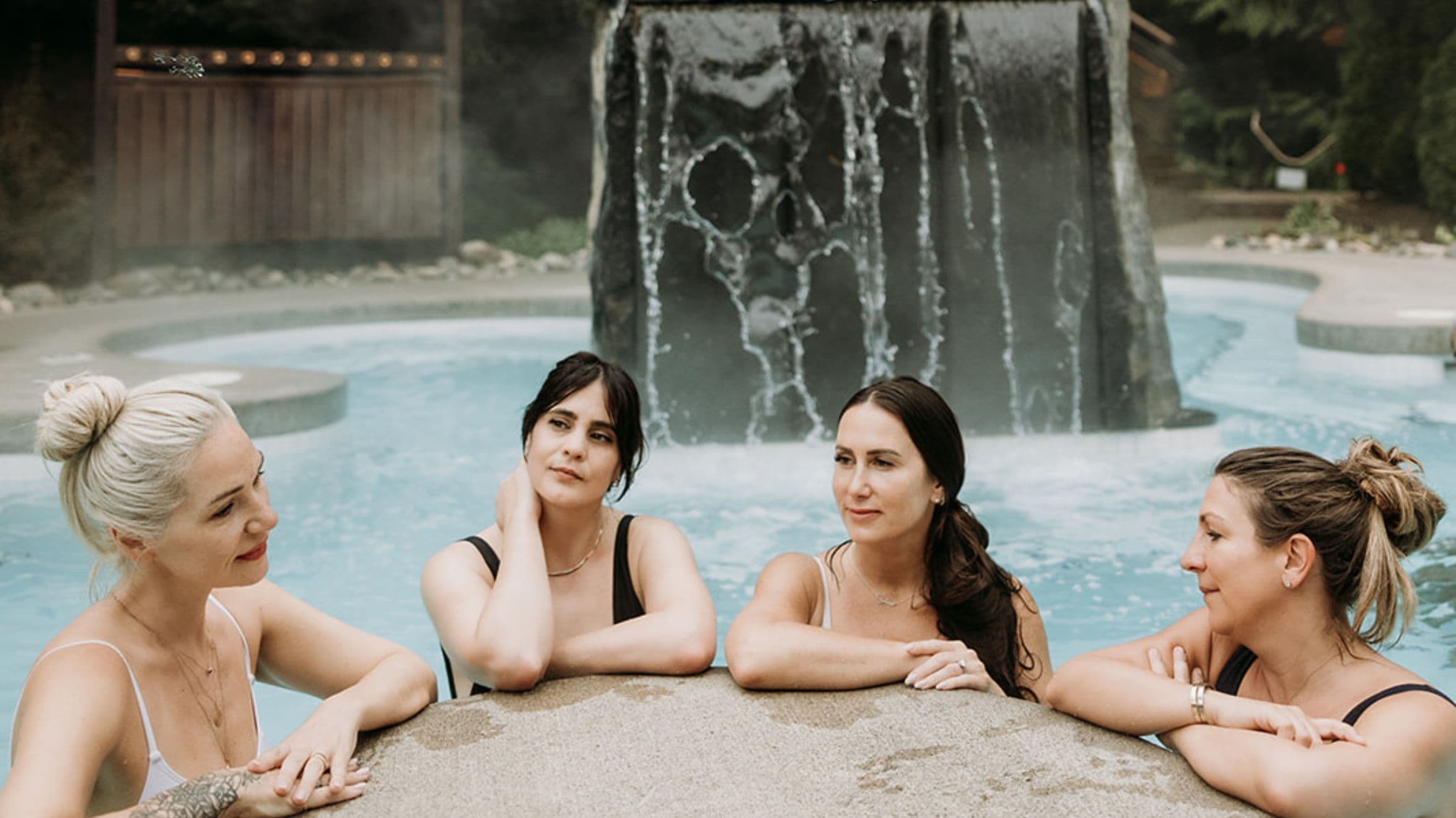 The women of Glimmer relax together in the hot pool at Scandinave Spa Whistler, thermal waterfall in the background.