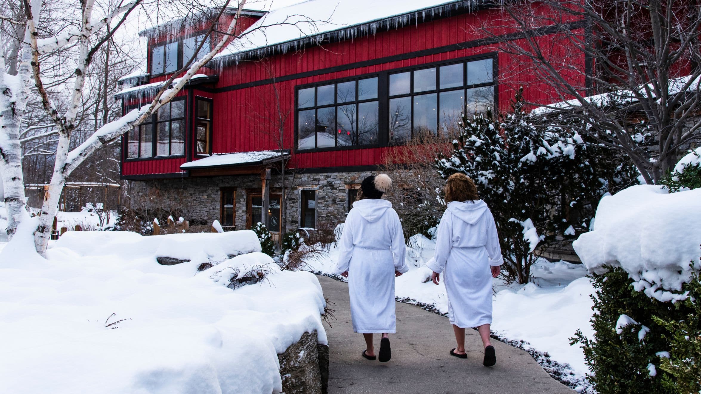 Two women wearing scandinave bathrobes are walking along a snow-lined path