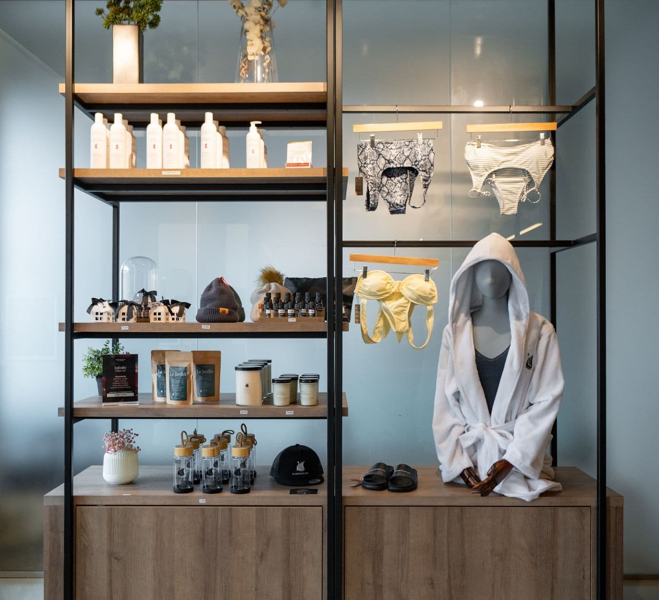 Étagères avec des items en boutique Scandinave Spa, dont les produits Carina Organics, bougies, maillots de bain pour hommes et femmes, sachets de tisane et mannequin en peignoir blanc.