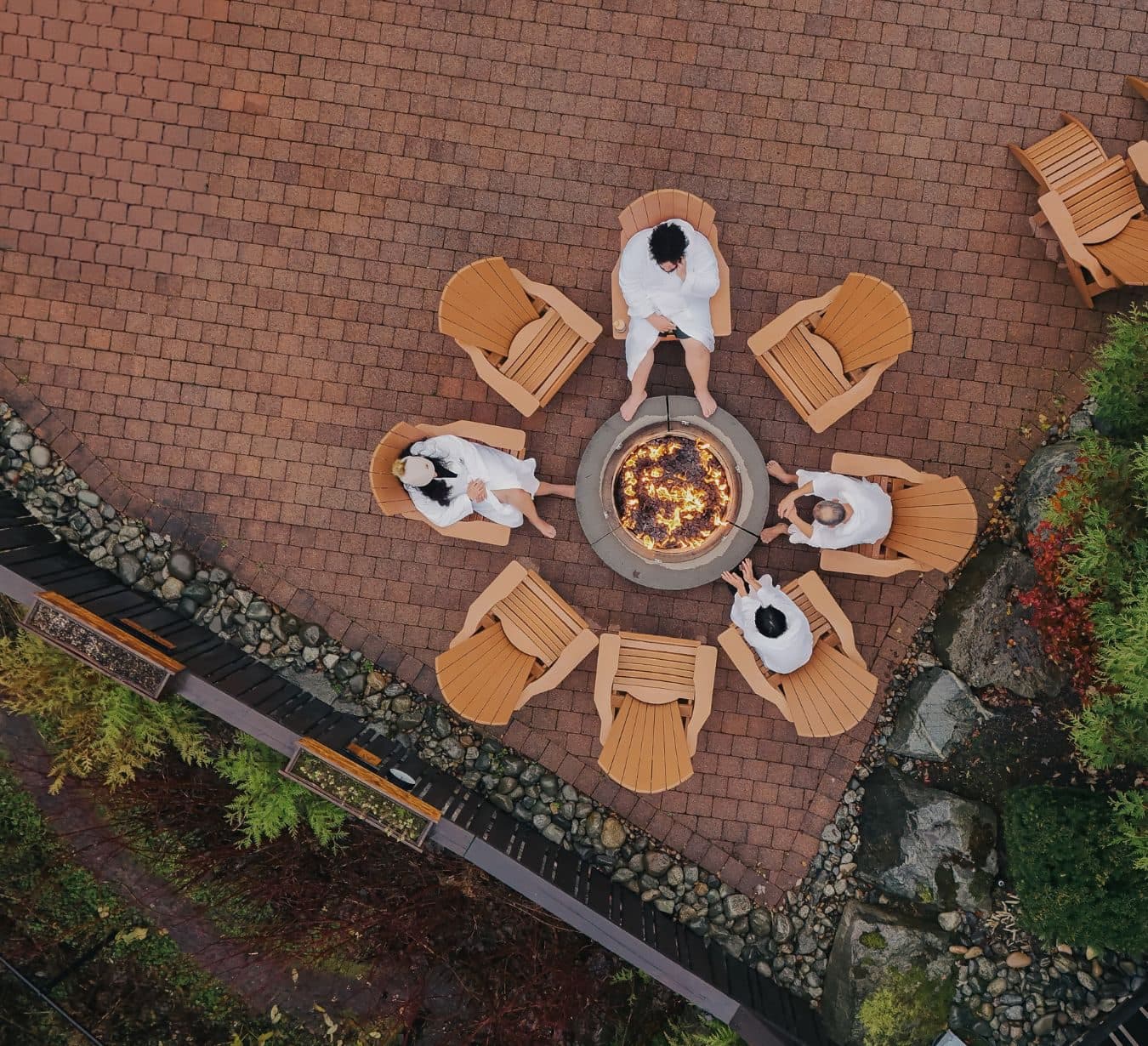Aerial view of people in white spa robes sitting around an outdoor fire pit in wooden Adirondack chairs.