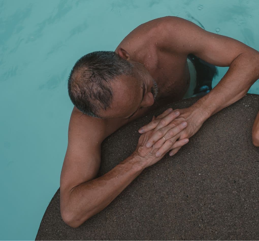 Man relaxing beside his partner in a hot pool, leaning forward with his hands folded on a smooth rock surface.