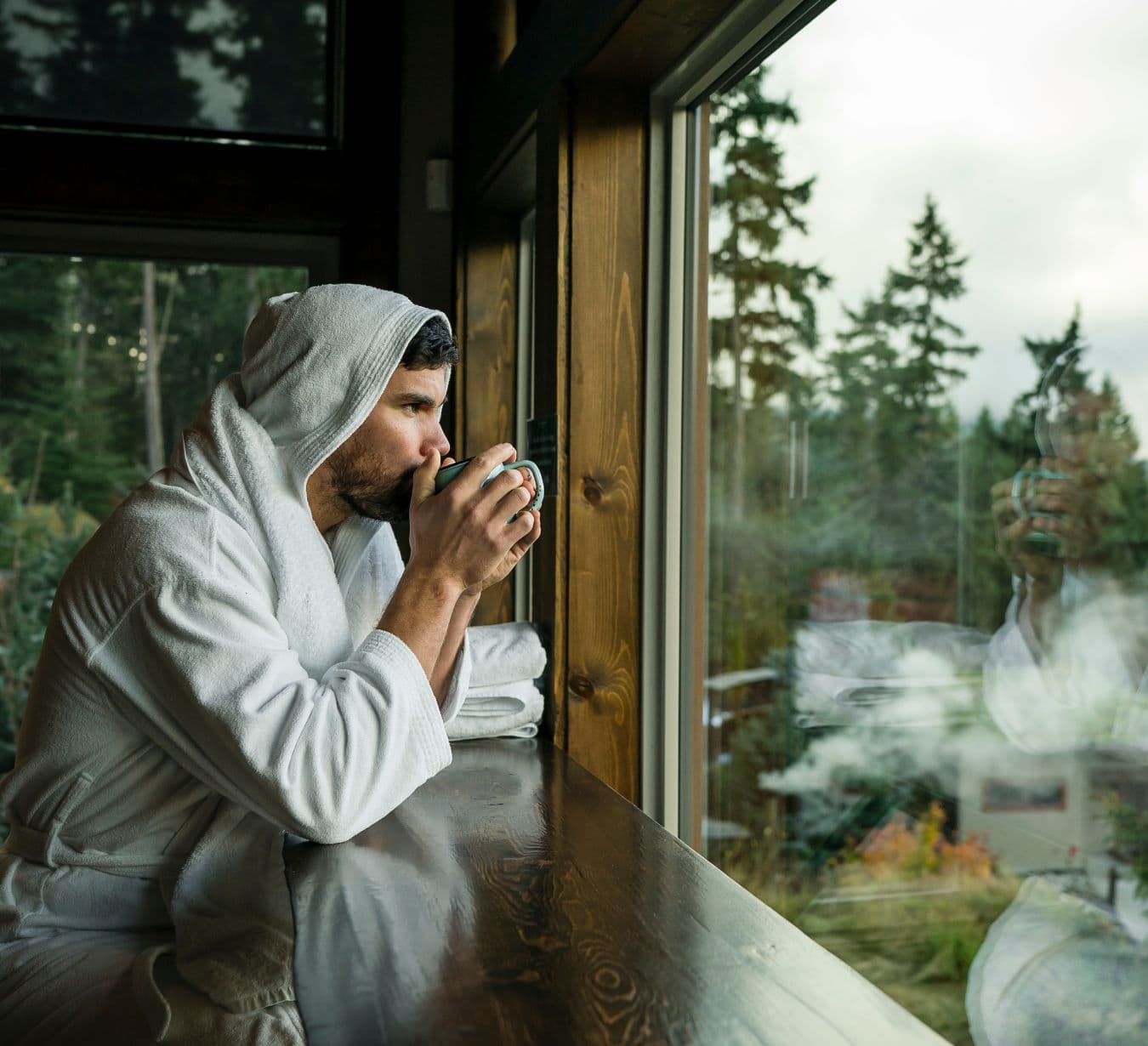 Person sitting in the bistro holding a warm drink while looking out a large window at the magnificent wooded landscape surrounding Scandinave Spa Whistler.