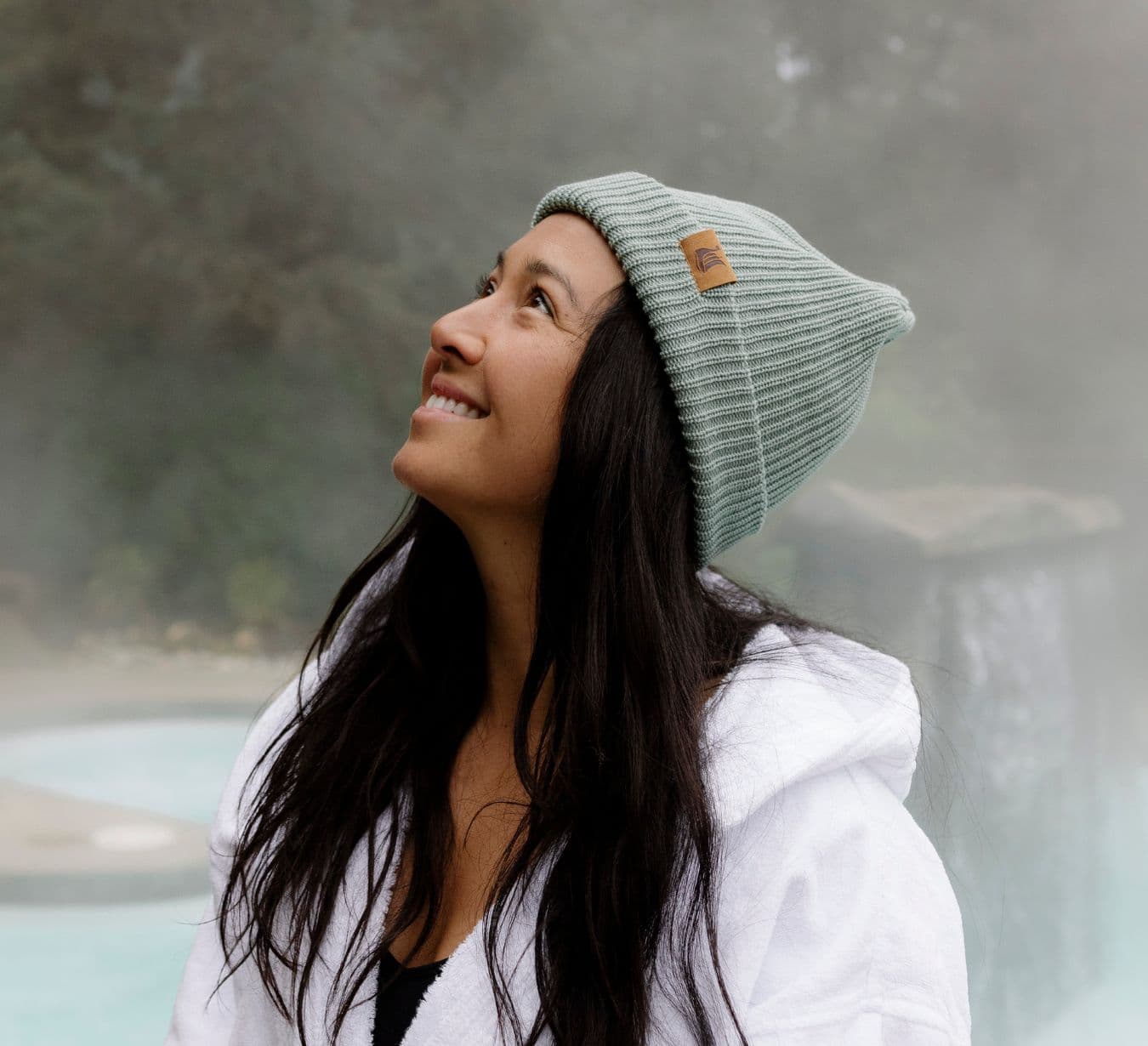 A smiling woman wearing a white bathrobe and a Scandinave Spa knit hat, standing near an outdoor hot tub surrounded by steam.